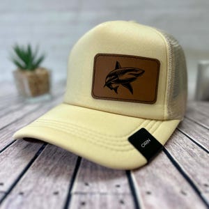 Può includere: Cappellino da camionista color crema con retro in rete e visiera curva. Una toppa in pelle marrone sul davanti presenta una grafica di squalo nero. Il cappello ha un'etichetta nera con la scritta "ORIN".