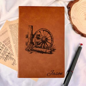 Peut inclure: Un journal en cuir marron avec un dessin rustique de roue de chariot et de poteau. Le nom "Jason" est inscrit en bas. Un stylet noir avec un embout rouge est à proximité.