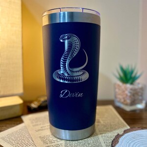 May include: A navy blue stainless steel travel mug featuring a silver cobra design and the name "Devin" engraved below. The mug has a silver rim and lid.