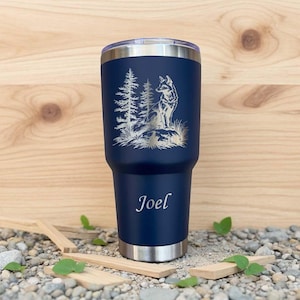May include: A navy blue insulated tumbler with a silver rim and lid. The tumbler features an engraved design of a wolf and pine trees. The name "Joel" is engraved below the design. The tumbler is designed to hold approximately 887 ml.