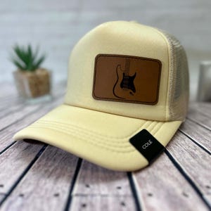 May include: Cream-coloured trucker hat with a brown leather patch featuring a black guitar outline. The hat has a mesh back and a curved brim. The tag on the brim reads "COLE".