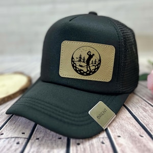 May include: Black trucker hat with a tan leather patch featuring a golf scene. The patch shows a golfer, trees, and a golf ball design. The hat has a curved brim and a mesh back. A silver tag with the name "BRENT" is attached.