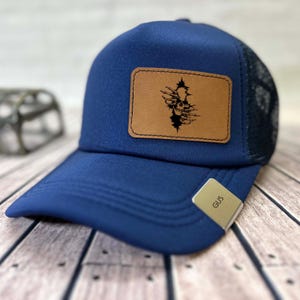May include: A navy blue trucker cap with a mesh back and a curved peak. It has a brown leather patch with a black skull design. A small tag on the brim reads "GUS".