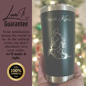 May include: A black and silver stainless steel tumbler with a personalized inscription that reads "Coach Kevin". The tumbler features a silver engraving of a person performing a grappling move.