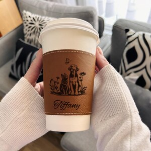 May include: A brown leather coffee cup sleeve featuring a design of a dog and cat sitting in a field of flowers. The sleeve is engraved with the name "Tiffany".