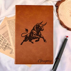 Peut inclure: Un journal en cuir marron avec une image en relief d'un taureau. Le nom "Gregory" est gravé en bas. Un stylet noir avec un embout rouge est posé à côté. Idéal pour l'écriture.