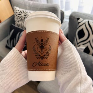 May include: A brown leather coffee cup sleeve with a fairy design and the name "Alicia" engraved on it.