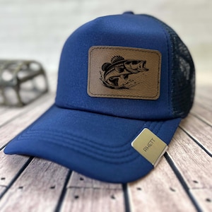May include: A navy blue trucker hat with a mesh back and a curved brim. It has a brown leather patch with a black graphic of a leaping fish. The tag on the brim reads "RHETT".