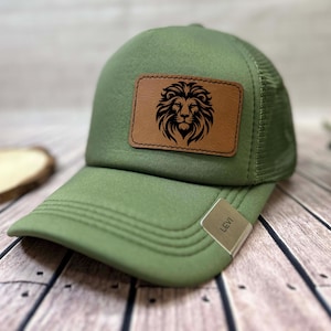 May include: A green trucker hat with a mesh back and a curved brim. A brown leather patch on the front features a black lion's head design. The hat has a tag that reads "LEVI".