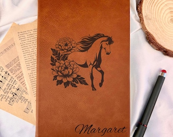 May include: A brown leather journal with a horse and floral design. The horse is etched in black, with the name "Margaret" in cursive below. A black stylus pen with a red tip rests nearby.