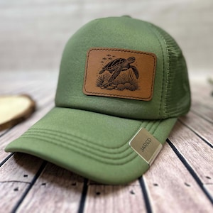 May include: Green trucker cap with a mesh back and a curved peak. A rectangular brown leather patch displays a detailed illustration of a sea turtle, coral, and fish. The tag on the peak reads "JARED".
