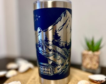 Könnte beinhalten: Ein Edelstahlbecher mit einem blauen und silbernen Design. Das Design zeigt eine Bergszene mit einem Fisch, der aus dem Wasser springt. Der Text "Gone Fishing" ist auf der Seite des Bechers aufgedruckt.