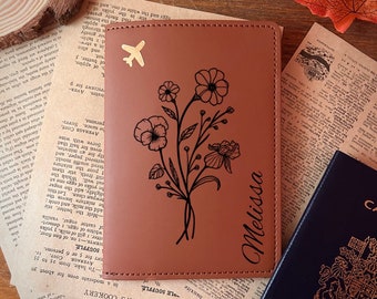 May include: A brown leather passport holder with a floral design and the name "Melissa" engraved on the front. The holder has a gold airplane charm.