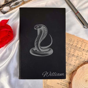 May include: Black leather journal featuring a silver cobra design and the name "William". The journal is rectangular with a textured surface. A pen and sheet music are in the background.