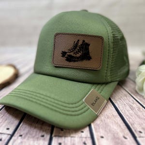May include: An olive green trucker cap with a brown leather patch showing a hiking boot design. The cap has a curved peak and a mesh back. A small tag on the brim reads "BLAKE".