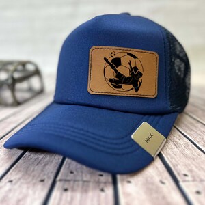 May include: A navy blue baseball cap with a mesh back and a curved brim. A rectangular, brown leather patch features a black silhouette of a soccer player kicking a ball. The tag on the brim reads "MAX".