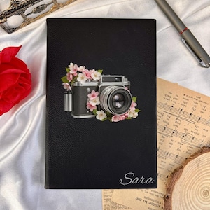 May include: A black rectangular notebook with a textured cover featuring a graphic of a vintage camera surrounded by pink cherry blossoms. The name Sara is printed in white script at the bottom right corner of the front cover.