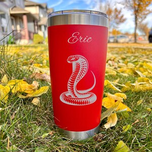 Peut inclure: Un gobelet rouge personnalisé avec un motif de cobra blanc et le nom "Eric" gravé sur le devant.