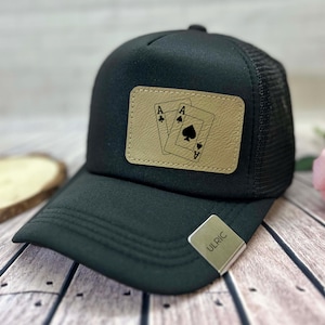 May include: Black trucker hat with a mesh back and a curved brim. A tan leather patch on the front displays two playing card aces. The hat has a snap closure and a metal tag that reads "ULRIC".