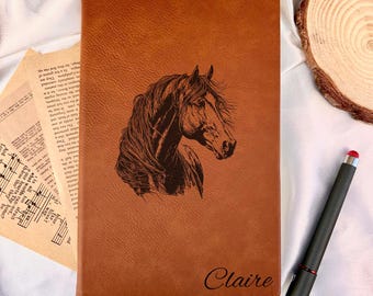 May include: A brown leather journal with a detailed horse head engraving. The name "Claire" is inscribed at the bottom. A black stylus pen with a red tip rests beside the journal.