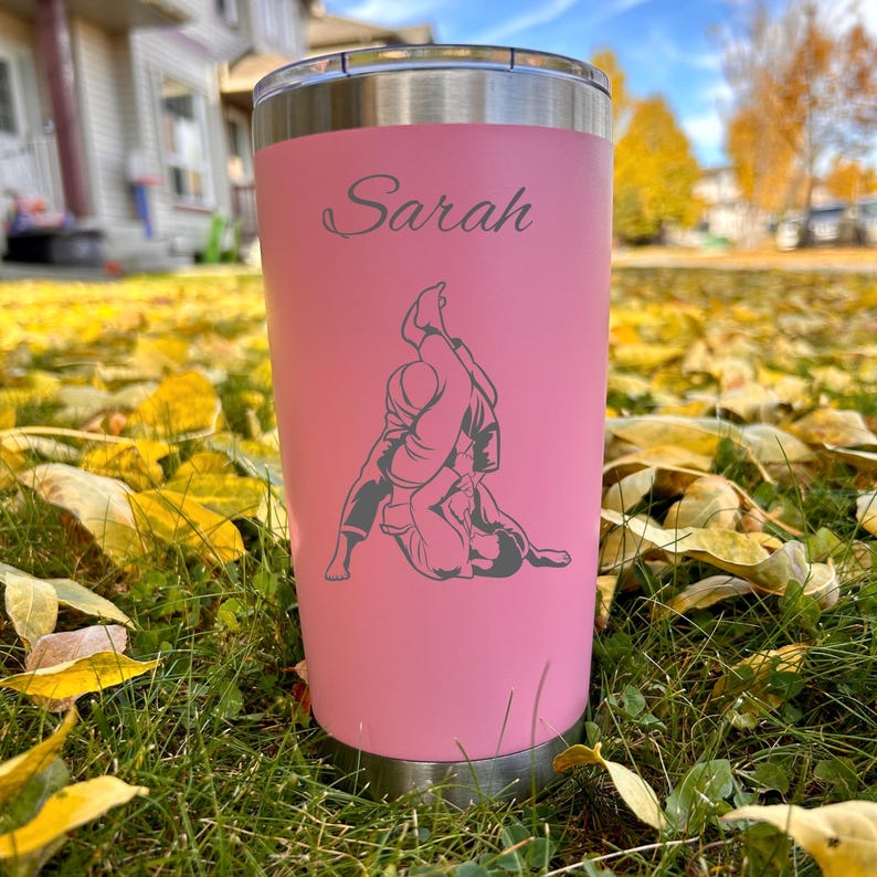 May include: Pink stainless steel tumbler with a black and white graphic of two people in a grappling position. The tumbler has the name "Sarah" engraved on the front.