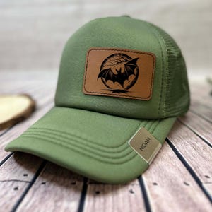 May include: Green baseball cap with a mesh back and a curved peak. The cap has a brown leather patch with a black bat design in front of a moon. The tag on the peak reads "NOAH".