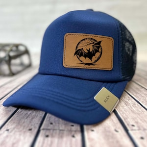 Può includere: Un cappellino da baseball blu navy con retro in rete e visiera curva. Il cappellino presenta una toppa in pelle marrone con una silhouette di pipistrello nero su un design di luna e albero. Un piccolo cartellino sulla visiera recita "ALEX".