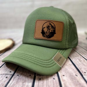 Pode incluir: Boné de caminhoneiro verde com um patch de couro castanho com uma cena de montanha. O boné tem uma parte traseira em malha e uma aba curva. Uma pequena etiqueta com o nome "Devin" está presa à aba.