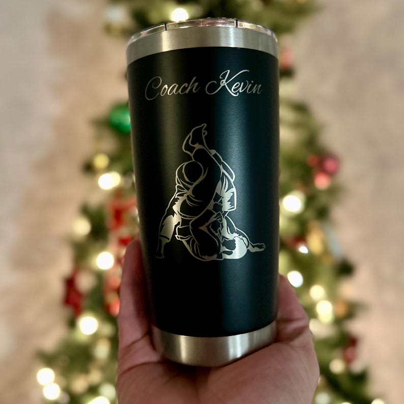 May include: A black stainless steel tumbler with a silver lid and base. The tumbler has an engraved image of two people in a wrestling hold and the text "Coach Kevin".