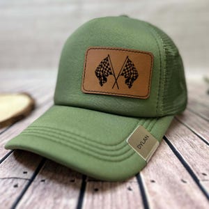 May include: Green trucker cap with a mesh back and a curved peak. A brown leather patch displays two crossed chequered flags. The tag on the peak reads "DYLAN".