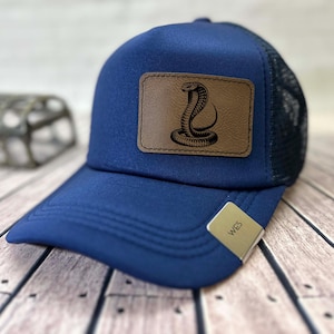 Peut inclure: Casquette de camionneur bleu marine avec un patch en cuir marron représentant un cobra détaillé. La casquette a un dos en filet et une visière incurvée. Une petite étiquette avec les lettres "WES" est attachée à la visière.