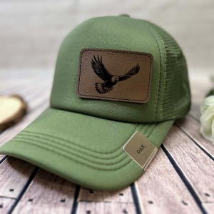 Puede incluir: Gorra de camionero verde oliva con un parche de cuero marrón con un diseño de águila negra. La gorra tiene una parte trasera de malla y una visera curva. Una pequeña etiqueta metálica en la visera dice "PAX".