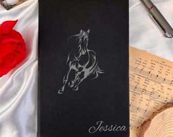 May include: Black leather journal with a running horse design and the name "Jessica" in cursive. The journal has a textured surface and a silver pen is visible in the background. A red rose and sheet music are also in the background.