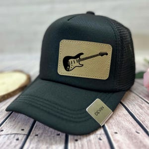 May include: Black trucker hat with a mesh back and a curved brim. A rectangular patch features a black guitar silhouette on a tan background. A small silver tag on the brim reads "Devin".