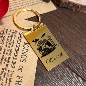 May include: Gold-colored keychain with a rectangular tag featuring a black and white illustration of a drum set. The tag is engraved with the text "ICINGS or FR", "CONFECTIONER'S ICING", and the name "Michael".