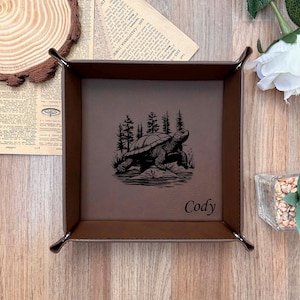May include: A brown leatherette valet tray featuring a black engraved turtle and forest design. The name "Cody" is printed in black. The tray is square with rounded corners and metal snap closures.