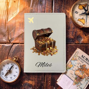 May include: A light gray passport holder featuring a graphic of an open wooden treasure chest overflowing with gold coins, pearls, and gemstones. A small yellow airplane icon is in the top left corner, and the name Miles is printed in black.