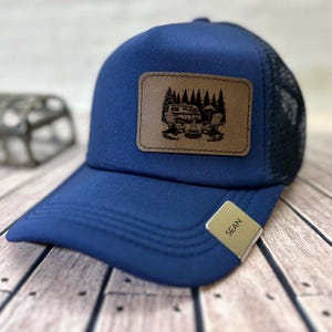May include: Navy blue trucker hat with a brown leather patch featuring a camping scene with a camper van, chairs, and a campfire. The hat has a mesh back and a small tag that says "SEAN".