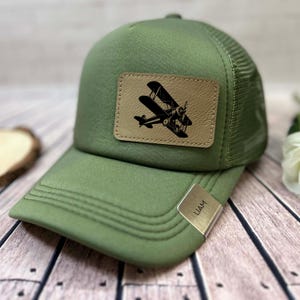 May include: Olive green trucker hat with a mesh back and a curved brim. A rectangular tan leather patch features a black biplane design. A silver metal tag on the brim reads "LIAM".