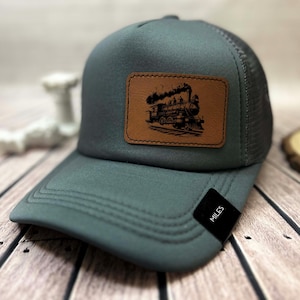 Könnte beinhalten: Graue Trucker-Cap mit einem braunen Lederpatch mit einer detaillierten Illustration einer Dampflokomotive. Die Kappe hat einen Mesh-Rücken und einen gebogenen Schirm. Ein kleines schwarzes Etikett auf dem Schirm trägt die Aufschrift "MILES".