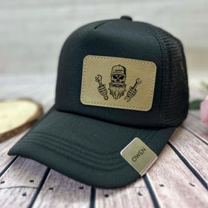 May include: Black trucker hat with a mesh back and a curved brim. A tan leather patch features a skull with a beard, holding wrenches, and the word "Garage" on the hat. A silver tag with the name "OWEN" is attached.