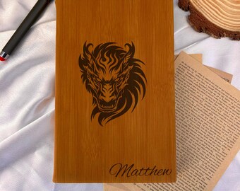 May include: A rectangular bamboo journal with a laser-etched dragon design and the name "Matthew" in a cursive font. The journal is a light brown color and has a smooth texture. A black pen rests on the white surface.