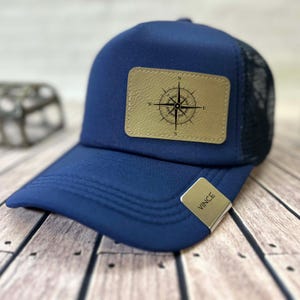 May include: A navy blue trucker hat with a black mesh back and a curved brim. The hat features a tan leather patch with a compass rose design. A small metal tag with the name "VINCE" is attached to the brim.