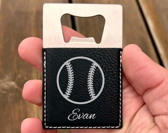 May include: A baseball-themed bottle opener with a stainless steel opener and a black leatherette case. The case features a white baseball graphic and the name "Evan" in script. White stitching outlines the case.