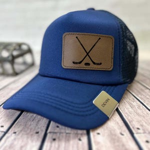 May include: A navy blue trucker cap with a brown leather patch displaying crossed hockey sticks and a puck. The cap has a mesh back and a curved peak. A small tag on the brim reads "Devin".