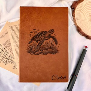 Puede incluir: Un diario de cuero marrón con un grabado detallado de una tortuga marina, corales y conchas. El nombre "Caleb" está inscrito en la parte inferior. Un lápiz táctil negro con punta roja está cerca. El diario es rectangular.
