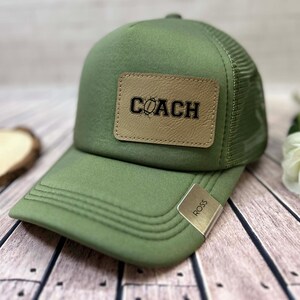 May include: An olive green trucker cap with a mesh back and a curved peak. The front features a tan leather patch with the word "COACH" in black. A silver tag with "ROSS" is on the side.