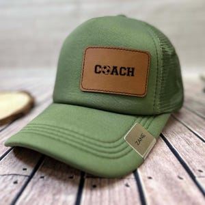 May include: Green baseball cap with a mesh back and a curved brim. A brown leather patch on the front displays the word "COACH" with a soccer ball graphic. The tag on the side reads "ZANE".