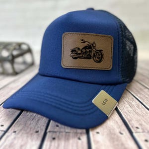 May include: A navy blue trucker hat with a brown leather patch featuring a motorcycle illustration. The hat has a mesh back and a curved brim. A small tag on the brim reads "LEVI".