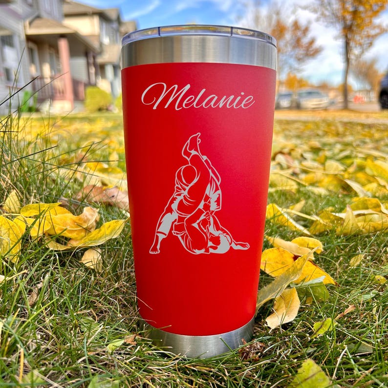 May include: Red stainless steel tumbler with a white silhouette of two people in a grappling position. The tumbler has the name "Melanie" engraved on the front.